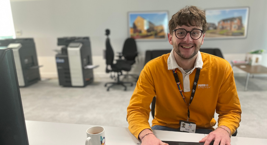 A member of staff wearing an orange jumper and smiling at their desk.